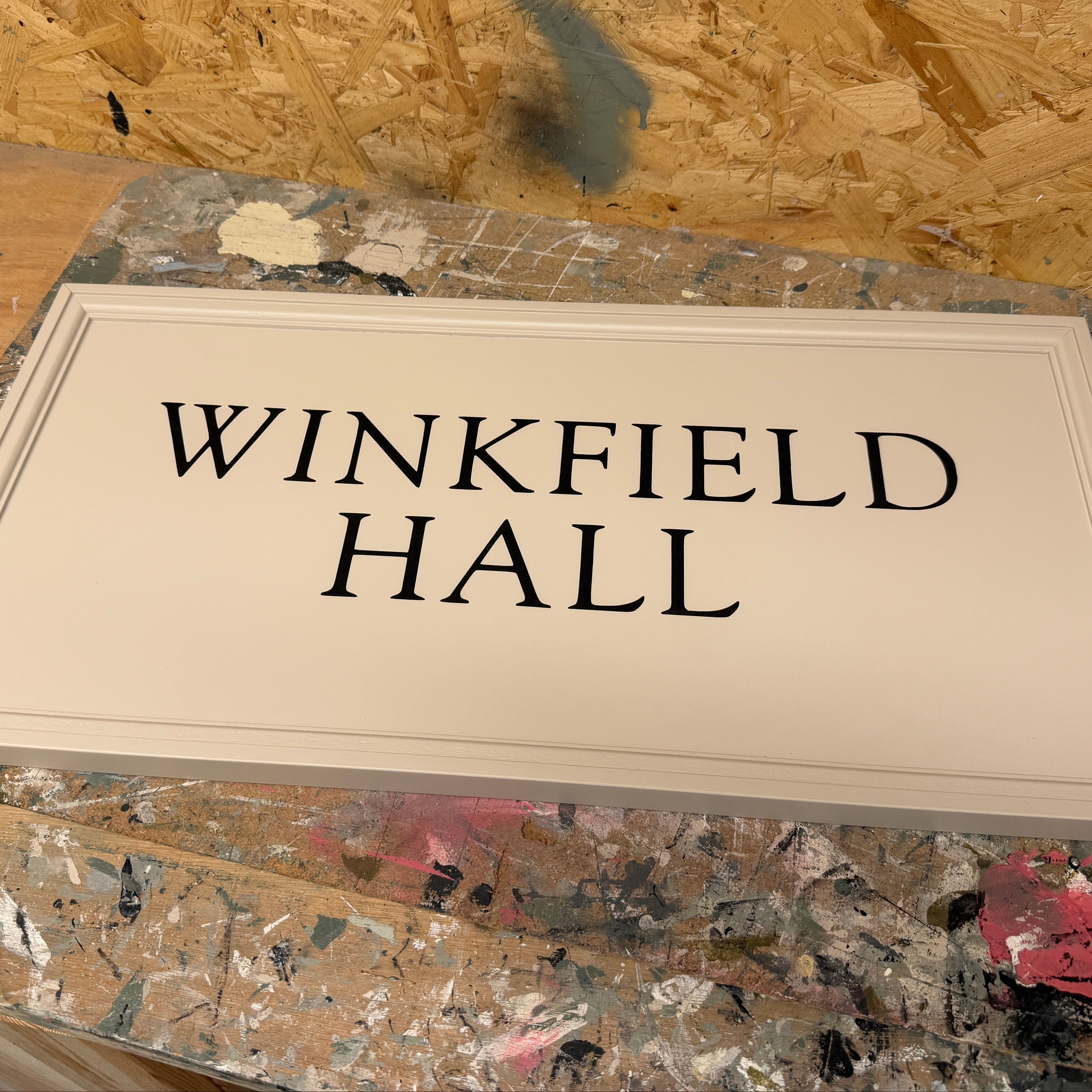 A rectangular cream-colored wooden sign with black text reading 'WINKFIELD HALL' displayed on a paint-splattered workbench.