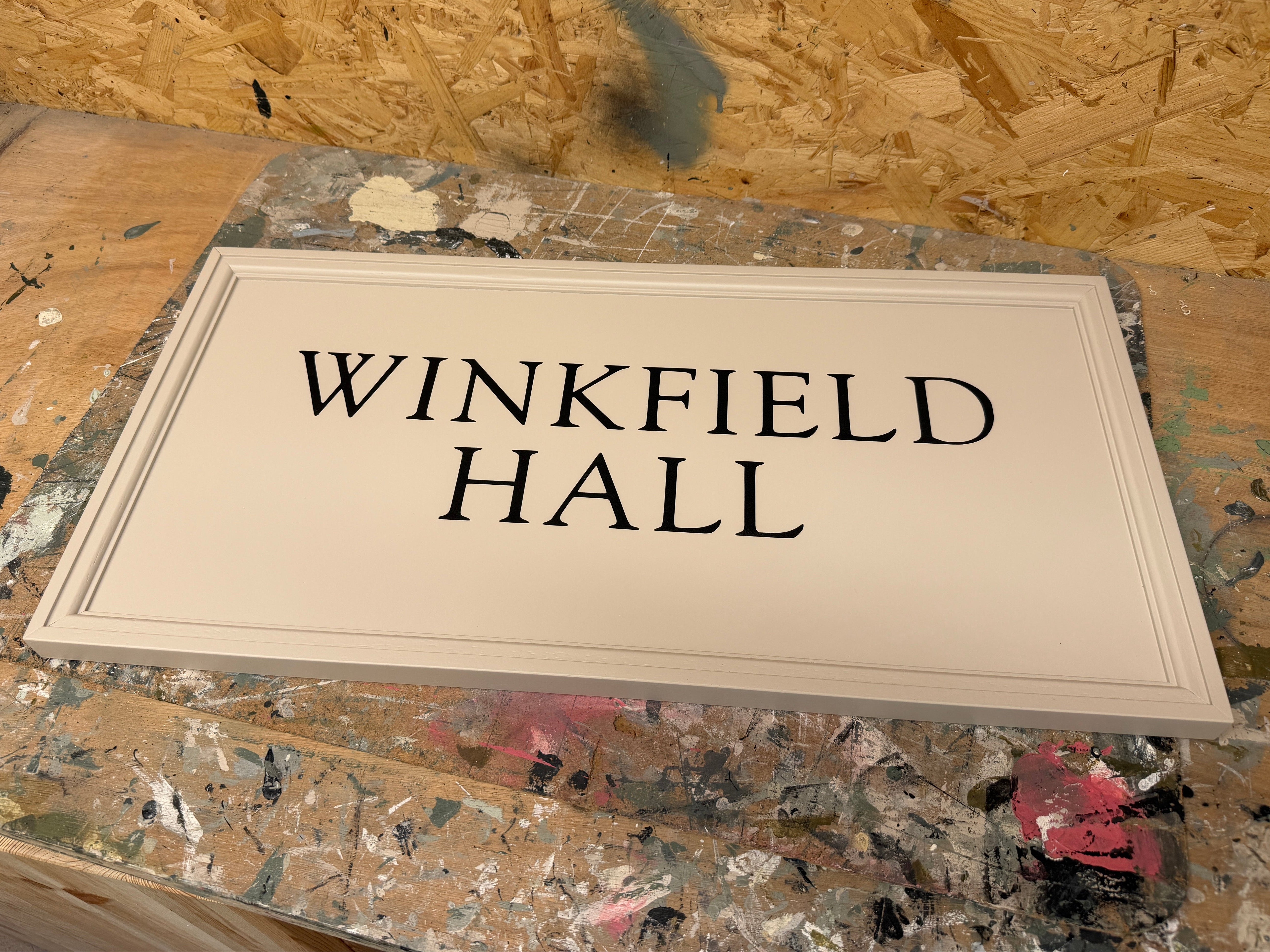 A rectangular cream-colored wooden sign with black text reading 'WINKFIELD HALL' displayed on a paint-splattered workbench.