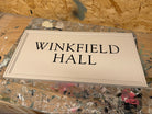 A rectangular cream-colored wooden sign with black text reading 'WINKFIELD HALL' displayed on a paint-splattered workbench.